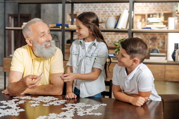 Mejor cooperación. Sonriendo feliz senior hombre y sus amados nietos haciendo un rompecabezas y uniendo tres piezas del rompecabezas