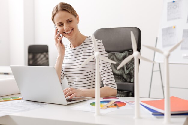 La mejor forma de comunicarse. Mujer profesional atractiva positiva sentada en la mesa y hablando por teléfono inteligente mientras diseña turbinas eólicas