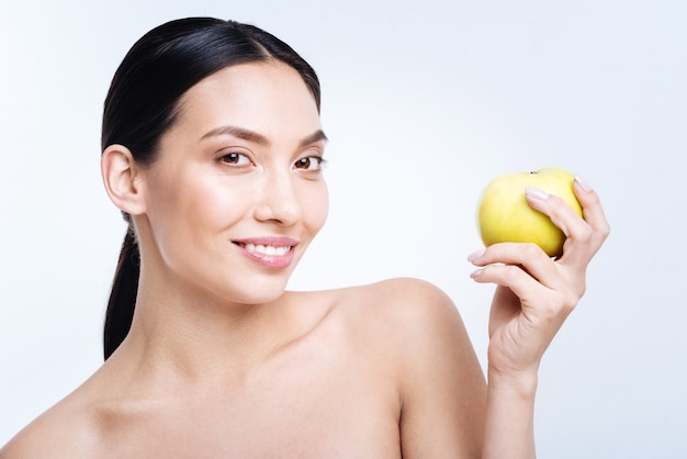 Mejor fruta. Encantadora mujer joven de pelo oscuro con hombros desnudos sosteniendo una manzana amarilla en sus manos y sonriendo mientras posa contra una pared blanca