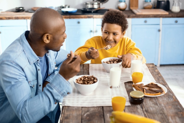 La mejor parte del día. Alegre niño preadolescente sentado en la mesa junto a su padre y comiendo cereales junto con él mientras intercambia sonrisas