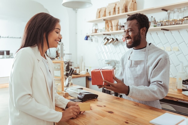 Mejor servicio. Alegre y agradable barista dando a su bonita clienta una caja con su pedido mientras la mujer sostiene una tarjeta de crédito, está lista para pagar