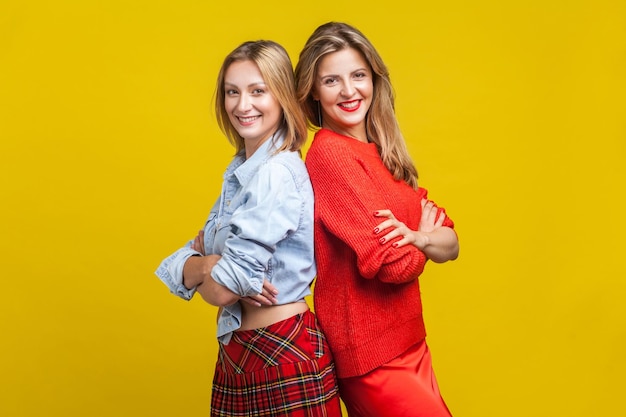 Las mejores amigas de pie de espaldas y con las manos cruzadas Retrato de dos mujeres encantadoras con ropa informal elegante sonriendo a la cámara en un estudio interior aislado en un fondo amarillo