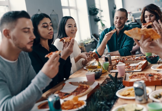 Entre mejores amigos. Grupo de jóvenes en ropa casual comiendo pizza y sonriendo mientras tienen una cena en el interior