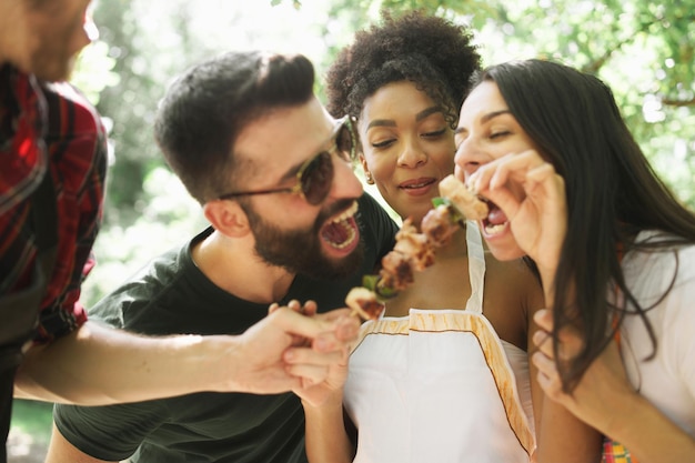 Mejores amigos multirraciales mordiendo brochetas de carne en el campo divirtiéndose juntos. Jóvenes en concepto de naturaleza