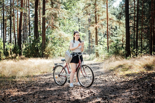 Las mejores aplicaciones de ciclismo Rastreador de bicicletas Mujer joven con mochila montando en bicicleta y mirando en el teléfono celular en el fondo del bosque de pinos Chica con bicicleta usando un teléfono enviando mensajes de texto en la aplicación de teléfono inteligente en un paseo por el bosque