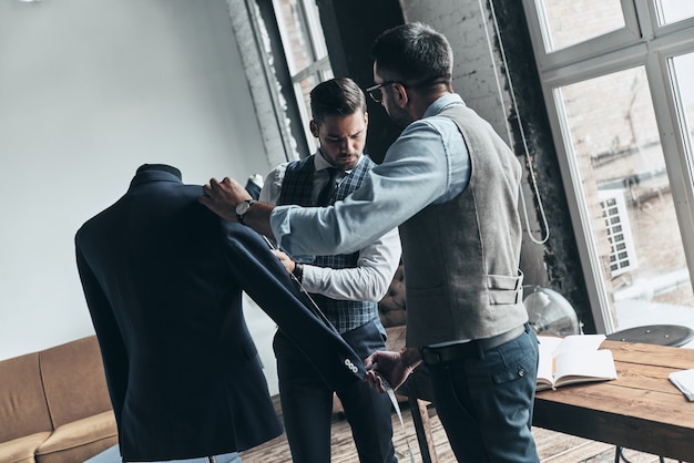 mejores diseños. Dos hombres jóvenes de moda midiendo la manga de las chaquetas mientras está de pie en el taller