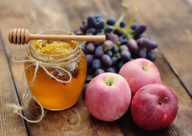 Mel em uma jarra bonita, eixo de colher de pau, uvas e maçãs numa superfície de madeira