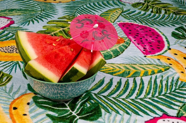 Melancia fatiada com um pequeno guarda-chuva vermelho, sobre um fundo tropical de folhas de palmeira.