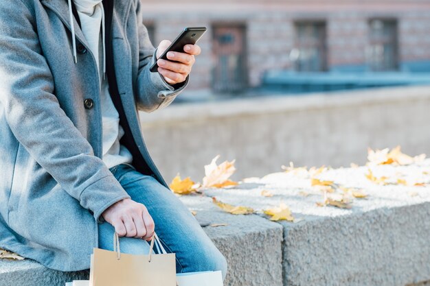 Melancolia de outono. Foto recortada de um homem sentado sozinho na parede com sacolas de compras, usando o smartphone.