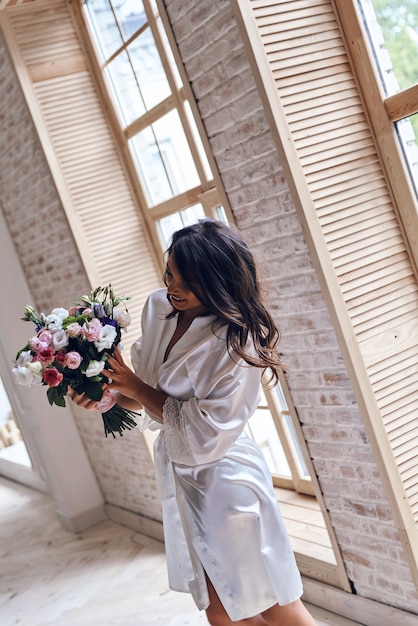 Melhor buquê de casamento. Mulher jovem e atraente em roupão de seda segurando um buquê e sorrindo em pé perto da janela