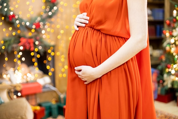 Melhor presente de Natal, mulher grávida sem rosto em um vestido vermelho abraça a barriga na sala de estar de Natal