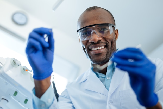 Melhor profissional. Alegre e agradável dentista masculino segurando um espelho bucal e uma sonda dentária e sorrindo para a câmera, sendo preparado para um exame de cárie