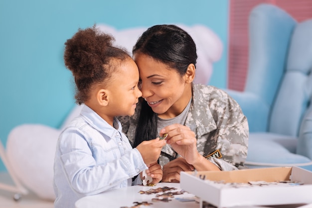 Melhor tempo. Linda e adorável mãe e filha maravilhosas compartilhando um momento fofo, abraçando enquanto está sentada à mesa branca e completando um quebra-cabeça