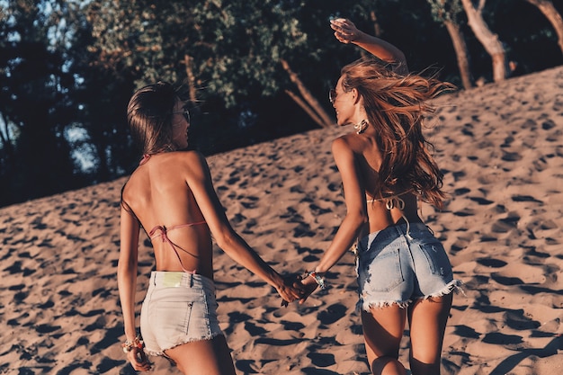 Melhores amigos. Duas mulheres jovens e atraentes em shorts e trajes de banho de mãos dadas enquanto correm na praia