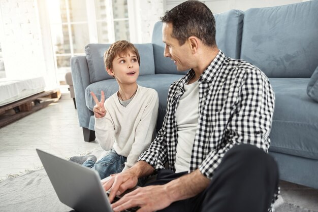 Foto melhores amigos. garoto louro bonito e alerta, sorrindo e sentado no chão com o pai e o pai segurando um laptop e se olhando