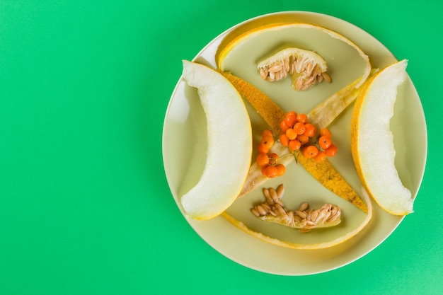 Melonenscheiben, Melonenkerne und -schalen auf einem grünen Teller. Melone und Vogelbeeren auf grünem Hintergrund. Vegetarische gesunde Ernährung. Draufsicht. Kopierraum
