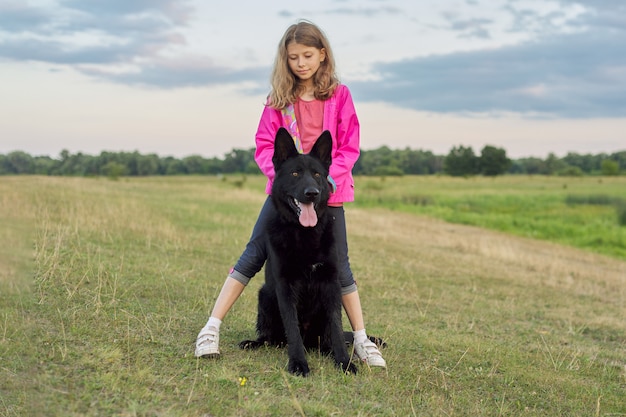 Menina abraçando grande cão preto pastor, criança e animal de estimação andando na natureza