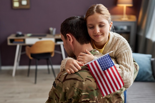 Menina adolescente abraçando o pai militar e segurando a bandeira americana em casa