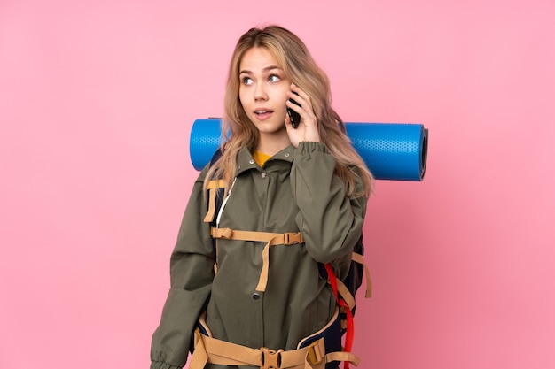 Menina adolescente alpinista com uma grande mochila isolada na rosa, conversando com alguém com o telefone celular