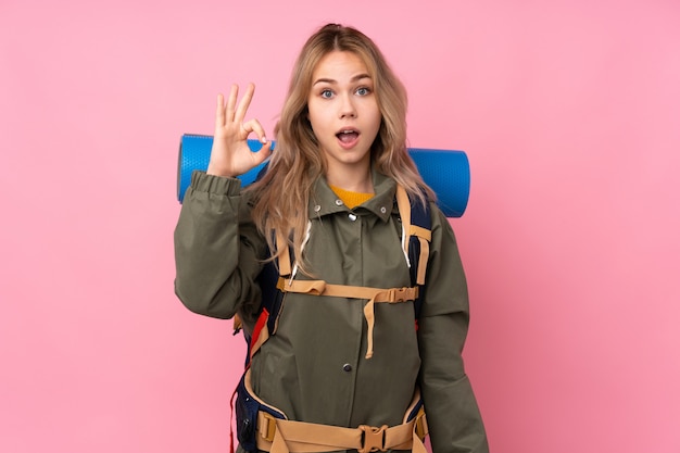 Menina adolescente alpinista com uma mochila grande no rosa surpreso e mostrando sinal ok