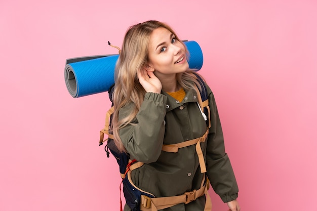 Menina adolescente alpinista russo com uma mochila grande na parede rosa ouvindo algo, colocando a mão na orelha