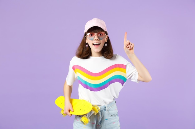 Menina adolescente animada em roupas vívidas, óculos com skate segurando o dedo indicador com grande nova ideia isolada em fundo violeta. Conceito de estilo de vida de emoções sinceras de pessoas. Mock up espaço de cópia.