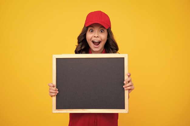 Menina adolescente animada em uniforme de construção vermelho e boné Trabalhador de adolescente segura quadro-negro isolado em fundo amarelo Conceito de renovação de crianças Copie o espaço no tabuleiro vazio mock up