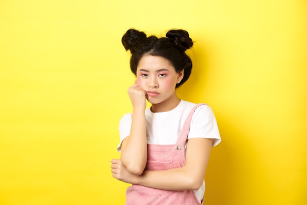 Menina adolescente asiática entediada com maquiagem elegante e roupas de verão, parecendo relutante e indiferente, parada no amarelo.