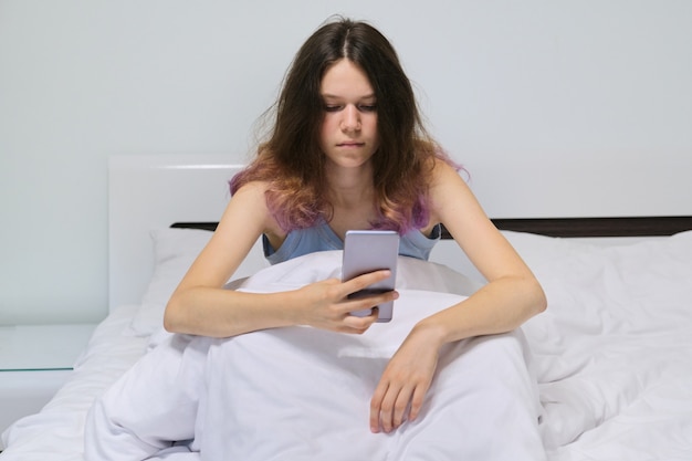 Menina adolescente cansada, sentado em casa na cama, olhando para o telefone móvel