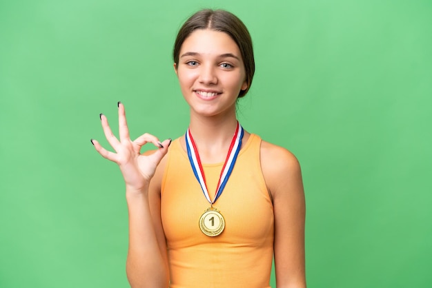 Menina adolescente caucasiana com medalhas sobre fundo isolado mostrando sinal de ok com os dedos
