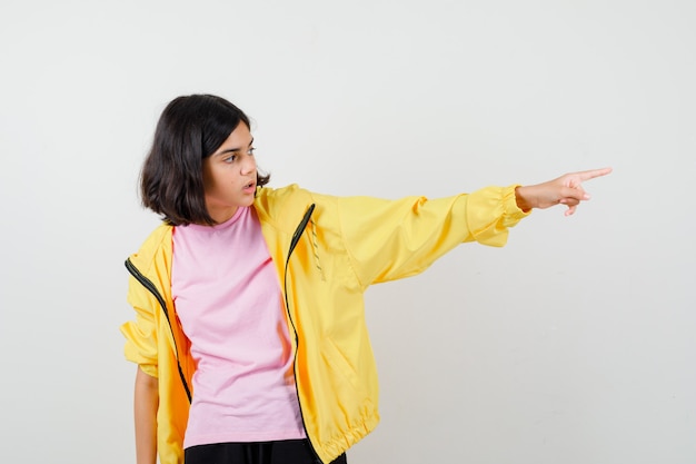 Menina adolescente com agasalho amarelo, camiseta apontando para o lado e parecendo surpresa, vista frontal.