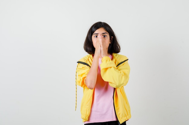 Menina adolescente com agasalho amarelo, camiseta de mãos dadas no rosto e parecendo assustada, vista frontal.