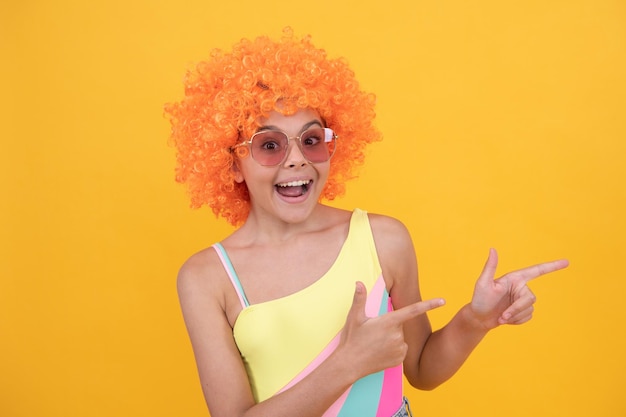 Menina adolescente com cabelo laranja ponto dedo verão praia moda anúncio dia da mentira feliz infância festa de aniversário criança espantada com peruca encaracolada se divertindo criança usar óculos escuros e maiô