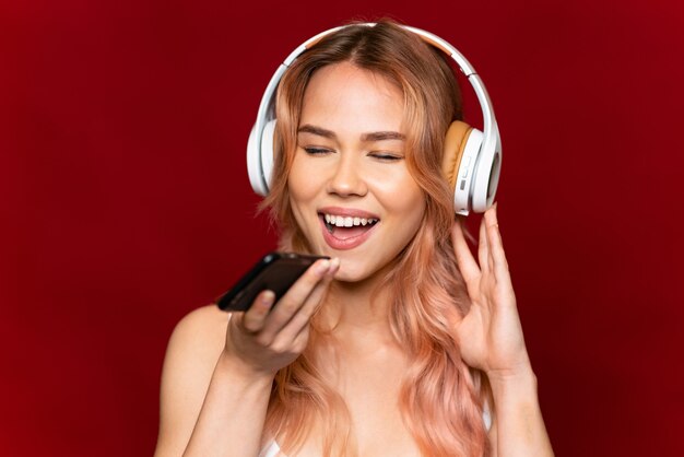 Menina adolescente com cabelo rosa em vermelho isolado ouvindo música com um celular e cantando