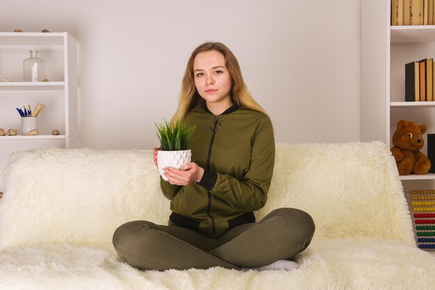 Menina adolescente com cara de acne sentada no sofá e segurando a imagem da planta de casa verde