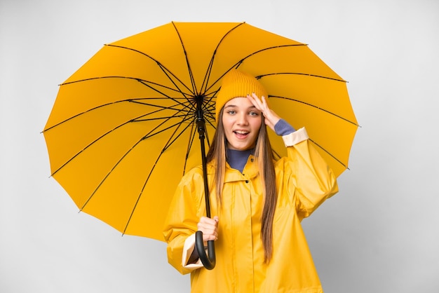 Menina adolescente com casaco à prova de chuva e guarda-chuva sobre fundo branco isolado com expressão de surpresa