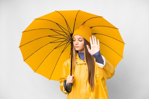 Menina adolescente com casaco à prova de chuva e guarda-chuva sobre fundo branco isolado, fazendo gesto de parada e decepcionada