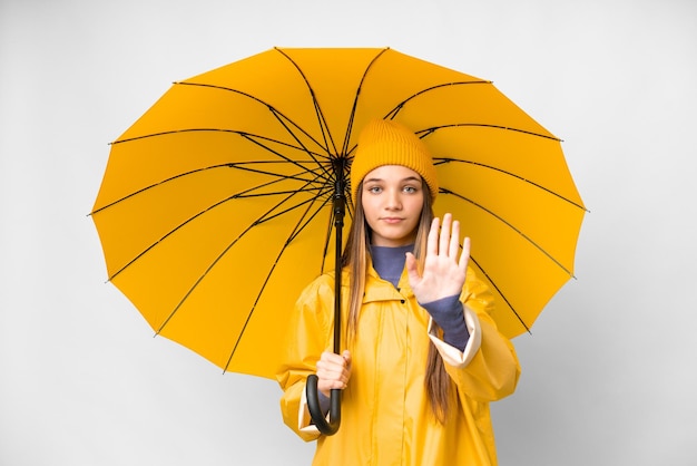 Menina adolescente com casaco à prova de chuva e guarda-chuva sobre fundo branco isolado, fazendo gesto de parada