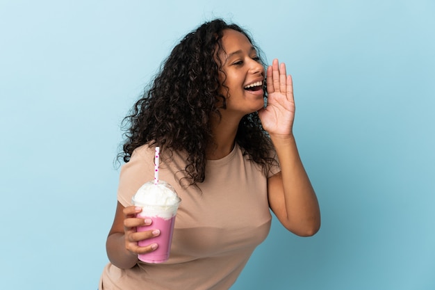 Menina adolescente com milkshake de morango isolada no azul gritando com a boca bem aberta para o lado