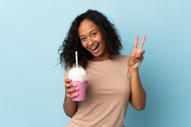 Menina adolescente com milkshake de morango isolada no azul sorrindo e mostrando sinal de vitória