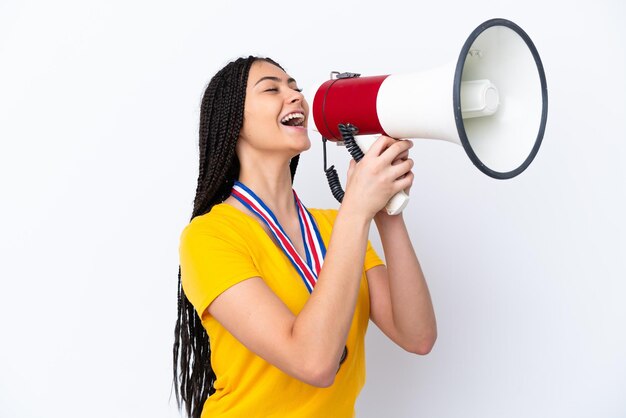 Menina adolescente com tranças e medalhas sobre fundo rosa isolado gritando através de um megafone