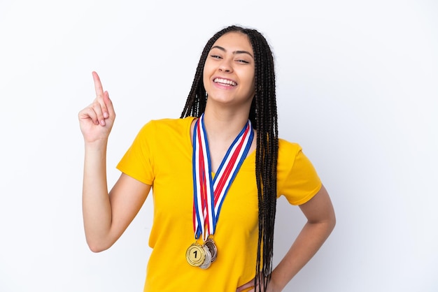 Menina adolescente com tranças e medalhas sobre fundo rosa isolado mostrando e levantando um dedo em sinal do melhor