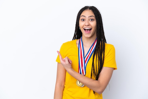 Menina adolescente com tranças e medalhas sobre fundo rosa isolado surpreso e apontando o lado