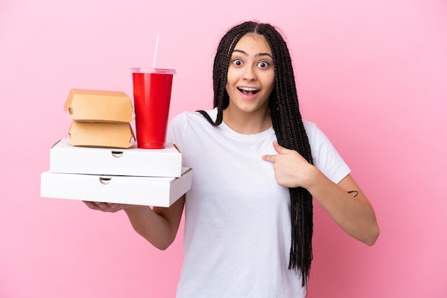 Menina adolescente com tranças segurando pizzas e hambúrgueres sobre fundo rosa isolado com expressão facial de surpresa