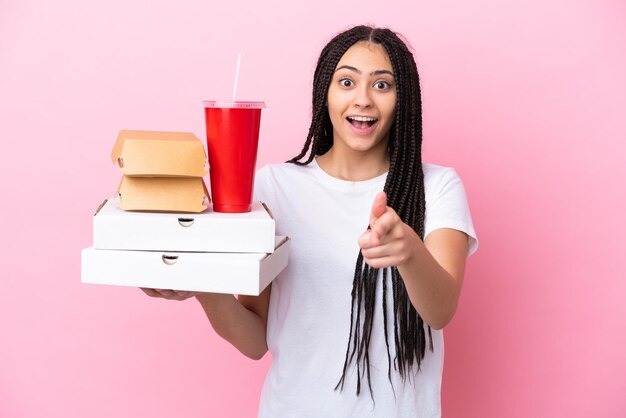 Menina adolescente com tranças segurando pizzas e hambúrgueres sobre fundo rosa isolado surpreso e apontando para a frente