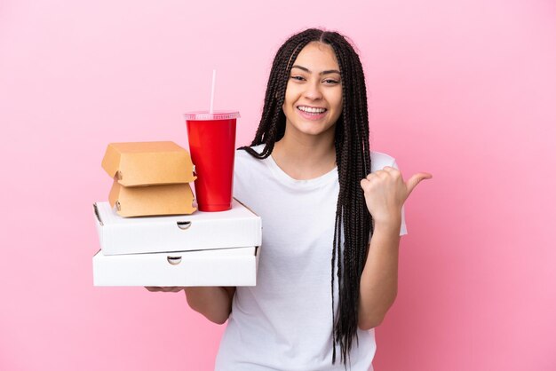 Menina adolescente com tranças segurando pizzas e hambúrgueres sobre um fundo rosa isolado apontando para o lado para apresentar um produto