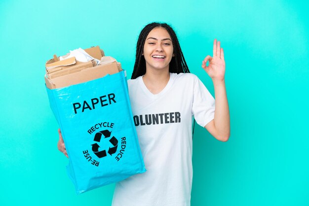 Menina adolescente com tranças segurando um saco para reciclar mostrando sinal de ok com os dedos