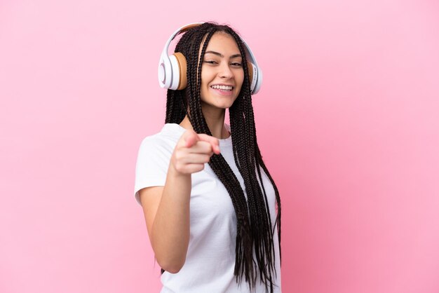 Menina adolescente com tranças sobre fundo rosa isolado ouvindo música e apontando para a frente