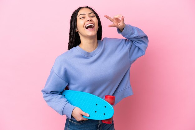 Menina adolescente com tranças sobre um fundo rosa isolado com um skate e uma expressão feliz