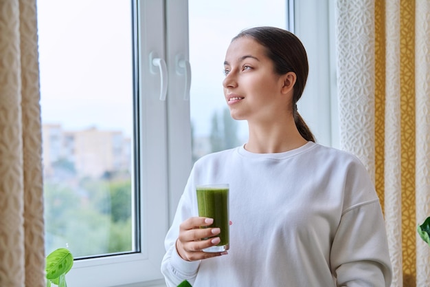 Menina adolescente com um copo de suco verde fresco pela manhã em casa perto da janela Comida saudável adolescência estilo de vida conceito de juventude